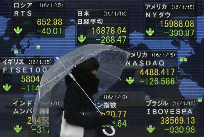 A pedestrian holding an umbrella walks past at an electronic board showing the stock market indices of various countries outside a brokerage in Tokyo